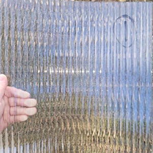 May include: A rectangular sheet of clear, textured glass featuring a repeating vertical ribbed pattern. The surface has a distinct linear design that distorts light and objects behind it. A circular mark is visible in the upper right corner.
