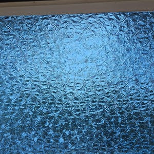 May include: A close-up of a textured, blue glass panel. The panel has a repeating pattern of stylized flower shapes, creating a shimmering effect. The glass is a vibrant shade of blue, with variations in tone across the surface.