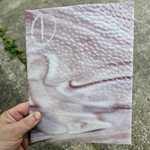 May include: A rectangular piece of stained glass with a swirling pattern of white and mauve. The top right corner has a textured, dotted design. A white oval with a vertical line is in the upper left corner. The glass is held by a hand.