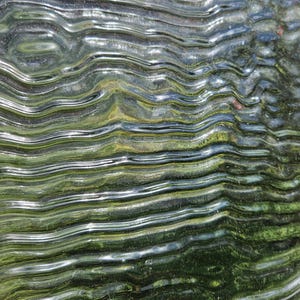 May include: Abstract image of a rippled glass surface. The glass has a wavy texture with horizontal lines, reflecting green and silver tones. The background shows hints of foliage.