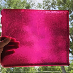 May include: A rectangular sheet of translucent fuchsia pink glass with a textured, rippled surface. The glass is held up against natural light, showing variations in color intensity and a semi-transparent quality across its flat, solid form.
