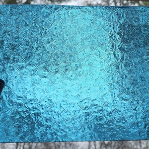 May include: A rectangular piece of textured, transparent, turquoise-colored glass. The glass has a repeating floral pattern. The glass is held up against a blurred background.