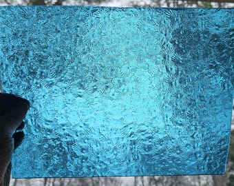 May include: A rectangular piece of textured, transparent, turquoise-colored glass. The glass has a repeating floral pattern. The glass is held up against a blurred background.