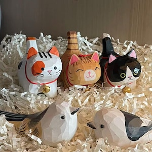 May include: A collection of hand-carved wooden figurines, including several cat figures with painted details and small bells, and two bird figures. The cats are in various colors, including white, orange, and black. The figurines are displayed on a bed of white paper.