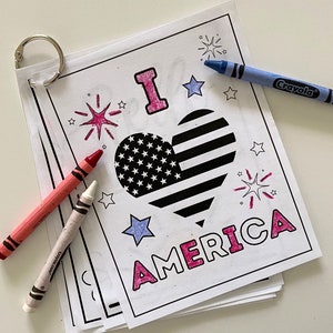 Op de afbeelding: Een kleurplaat met de woorden "I love America" in roze letters. In het midden van de pagina staat een hartvormige Amerikaanse vlag. Er zijn sterren en vuurwerk rond het hart. Crayola-kleurpotloden liggen op de pagina.