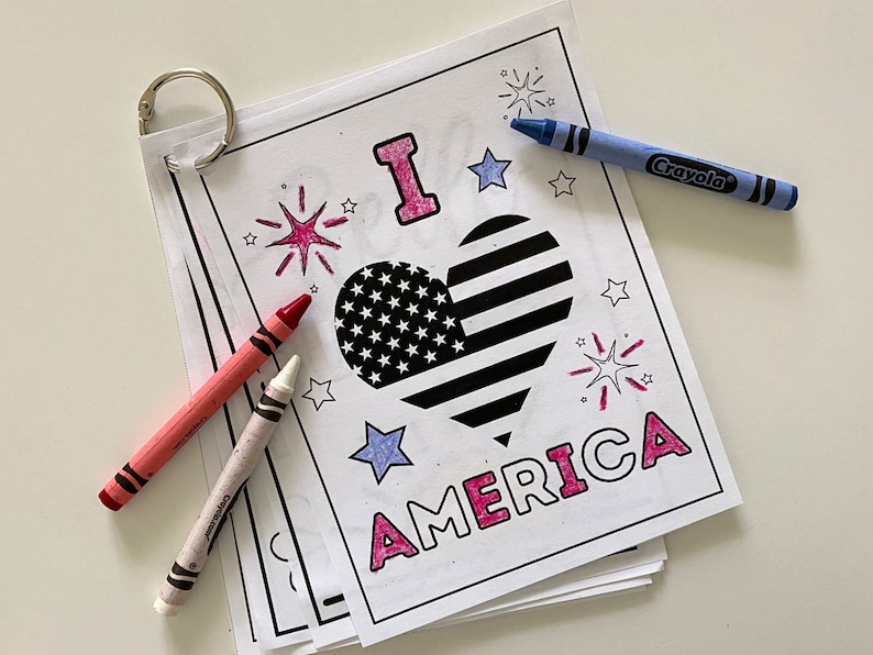 Op de afbeelding: Een kleurplaat met de woorden "I love America" in roze letters. In het midden van de pagina staat een hartvormige Amerikaanse vlag. Er zijn sterren en vuurwerk rond het hart. Crayola-kleurpotloden liggen op de pagina.