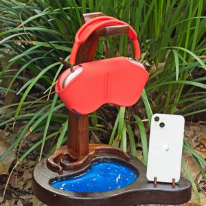 Puede incluir: Un soporte de madera con una incrustación de resina azul, un soporte para teléfono y un soporte para auriculares rojos.