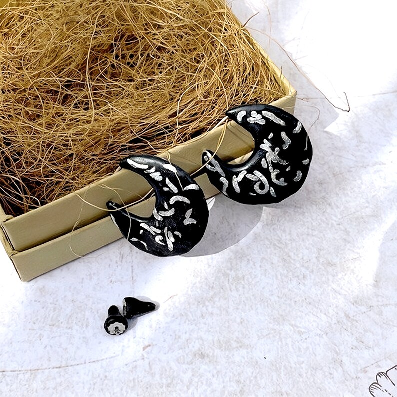 May include: Black crescent-shaped earrings with silver abstract designs. The earrings are displayed on a white surface, near a small box filled with brown straw. A pair of matching earring backs are also visible.