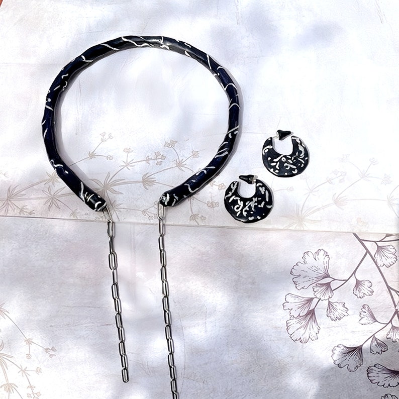 May include: A black and white necklace and earring set. The necklace is a thick, circular band with a chain. The earrings are half-circle shaped with a matching black and white design. The jewelry set is displayed on a white surface.