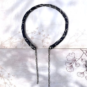 May include: A black necklace with a white, swirling pattern. The necklace has a circular design with two silver chains hanging down. The background is a light, floral pattern.