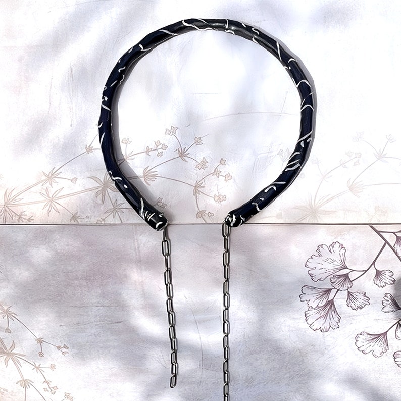 May include: A black necklace with a white, swirling pattern. The necklace has a circular design with two silver chains hanging down. The background is a light, floral pattern.