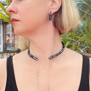 May include: A black choker necklace with a silver and white pattern, paired with matching hoop earrings. The jewelry is worn with a black top. The necklace has a silver chain that hangs down.