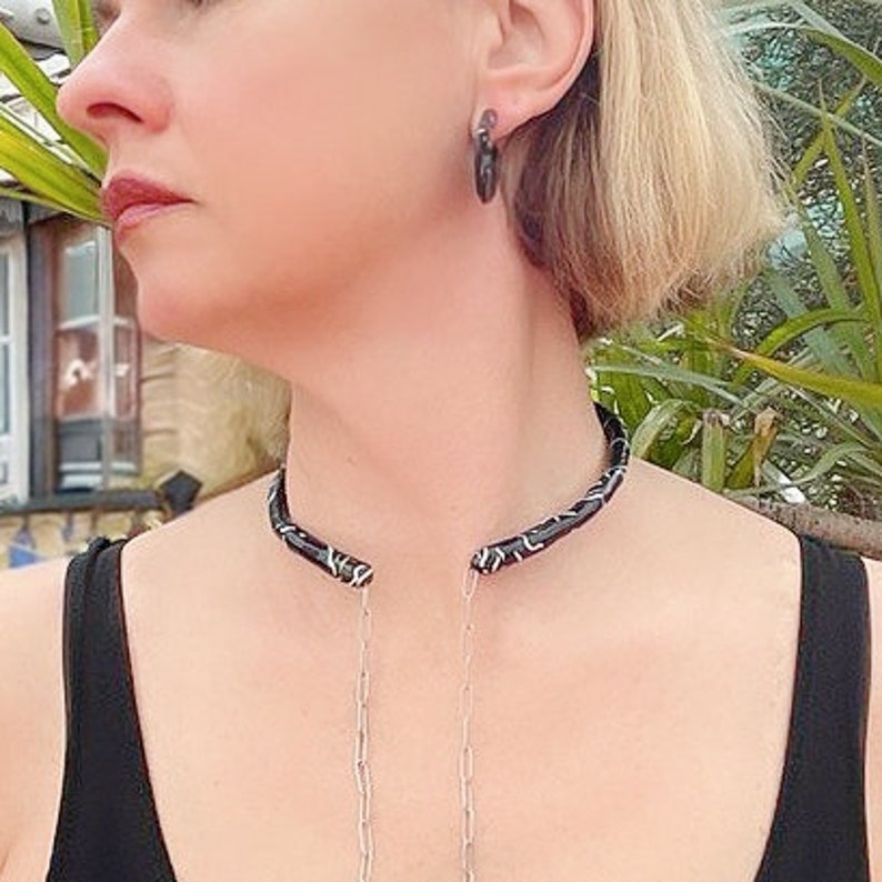 May include: A black choker necklace with a silver and white pattern, paired with matching hoop earrings. The jewelry is worn with a black top. The necklace has a silver chain that hangs down.