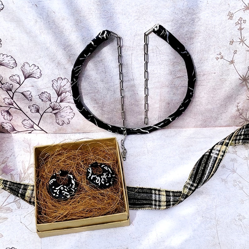 May include: A set of black and white earrings and a matching necklace. The earrings are in a small box filled with straw. The necklace and earrings have a black base with white decorative patterns. A black and white plaid ribbon is in the background.
