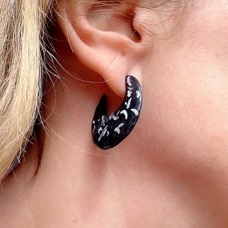 May include: A black crescent-shaped earring with white abstract designs is worn on an ear. The earring is a dark color with a curved shape, and the white designs add contrast. The earring is a unique piece of jewelry.