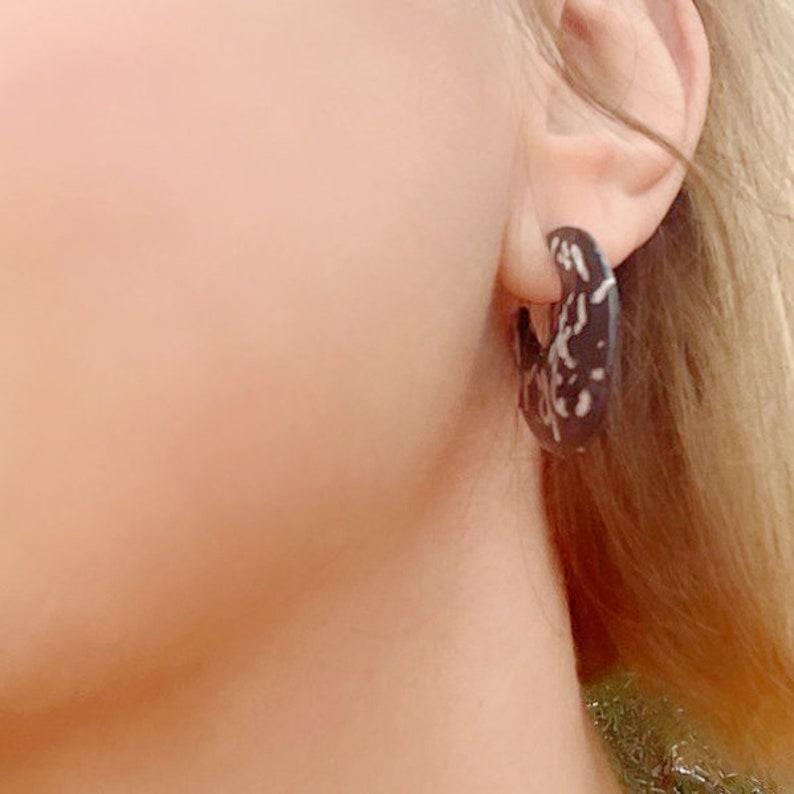 May include: A close-up of a person's ear wearing a dark brown hoop earring. The earring has a white, speckled pattern. The person has blonde hair and fair skin. The earring is a bold, modern accessory.