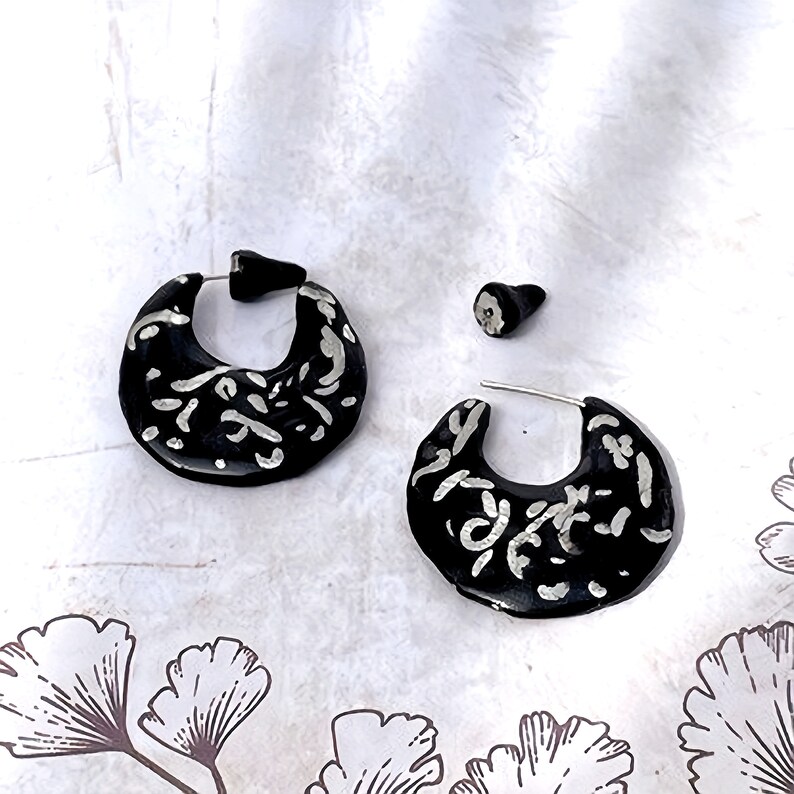 May include: Black and silver earrings with a unique design. The earrings are a crescent shape with a black base and silver swirls. The earring posts are silver. The earrings are displayed on a white surface.