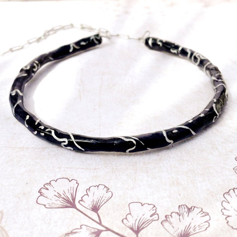 May include: A black choker necklace with a silver chain and decorative silver swirls. The necklace is made of a dark, textured material and has a simple, elegant design. The necklace is approximately 14 inches in length.