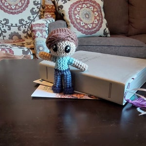 May include: A handmade crochet doll with a brown cap, light blue shirt, and blue pants. The doll has large black eyes and outstretched arms. It is standing on a book. The doll is a unique, handcrafted item.
