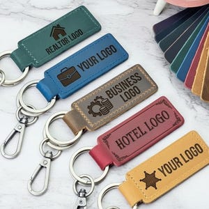 May include: Five rectangular leather keychains with metal lobster clasps and split rings. The keychains are teal, blue, brown, red, and yellow, each featuring custom engraved text and icons including REALTOR LOGO, YOUR LOGO, BUSINESS LOGO, HOTEL LOGO, and YOUR LOGO.