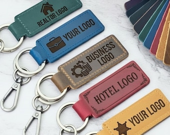 May include: Five rectangular leather keychains with metal lobster clasps and split rings. The keychains are teal, blue, brown, red, and yellow, each featuring custom engraved text and icons including REALTOR LOGO, YOUR LOGO, BUSINESS LOGO, HOTEL LOGO, and YOUR LOGO.