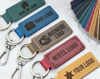 May include: Five rectangular leather keychains with metal lobster clasps and rings. Colors include teal, blue, brown, maroon, and tan. Each keychain features engraved text and icons: REALTOR LOGO, YOUR LOGO with a briefcase, BUSINESS LOGO, HOTEL LOGO, and YOUR LOGO with a star.