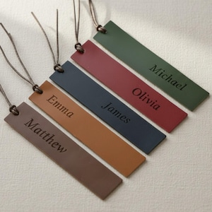 May include: Five rectangular bookmarks in brown, tan, navy blue, red, and olive green. Each bookmark features a thin leather cord at the top and is engraved with a name: Matthew, Emma, James, Olivia, and Michael in a serif font style.