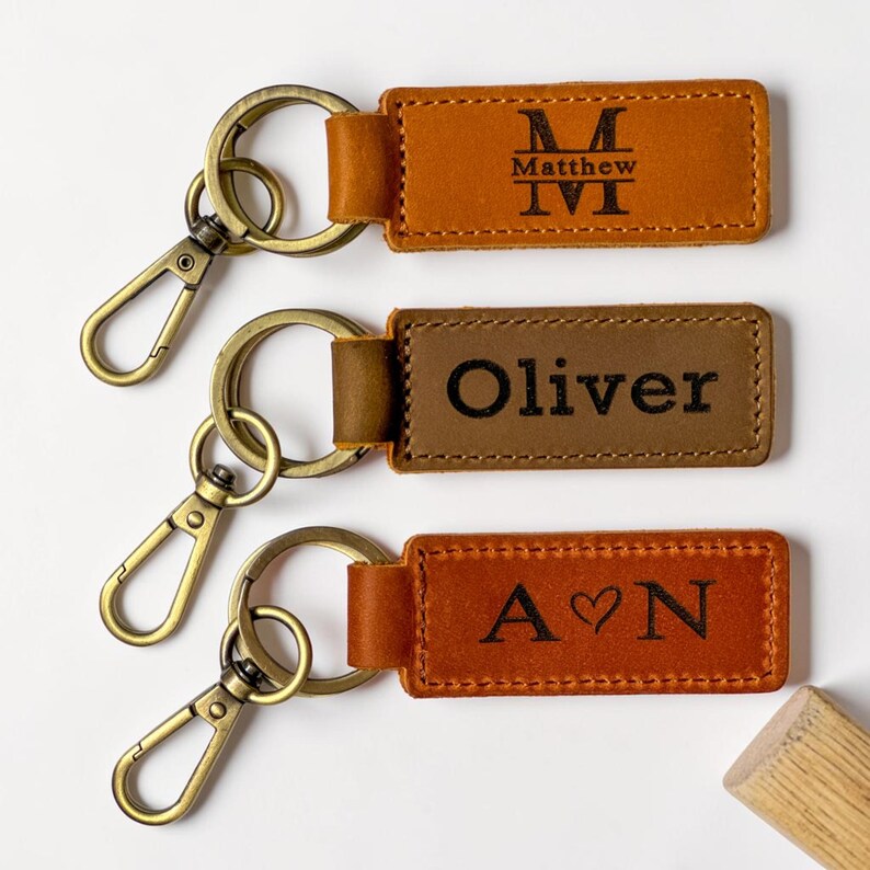 May include: Three rectangular leather keychains with antique brass-toned metal lobster clasps and split rings. The top keychain is tan with the name Matthew and a monogram. The middle is brown with the name Oliver. The bottom is tan with A heart N.