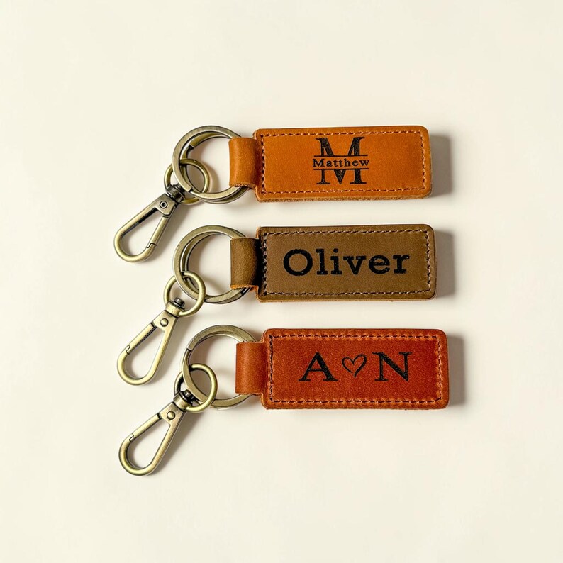 May include: Three rectangular leather keychains with bronze-toned metal lobster clasps and rings. The top keychain is tan with the name Matthew and a large M. The middle is brown with the name Oliver. The bottom is reddish-brown with A heart N.