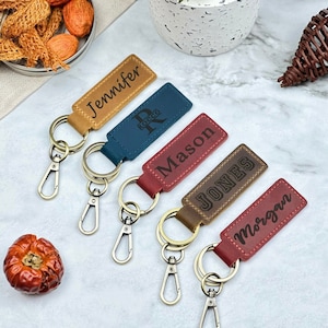 May include: Five personalized leather keychains with metal rings and clips. The keychains are in different colors, including brown, red, blue, and tan. The names "Jennifer", "Rocco", "Mason", "Jones", and "Morgan" are engraved on the keychains.