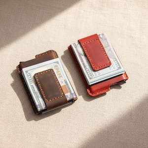 May include: Two leather money clips, one brown and one red, each holding a stack of US hundred-dollar bills. The clips have a magnetic closure and are stitched with a contrasting thread. The wallets are displayed on a neutral-colored surface.