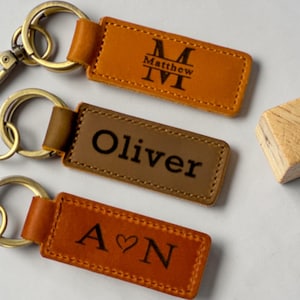 May include: Three rectangular leather keychains with brass-toned metal rings. The top keychain is tan with the name Matthew and a monogram. The middle keychain is brown with the name Oliver. The bottom keychain is tan with the letters A and N separated by a heart.