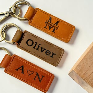 May include: Three rectangular leather keychains with metal rings and clasps. The top keychain is tan with a monogram and the name Matthew. The middle keychain is brown with the name Oliver. The bottom keychain is tan with the letters A and N and a heart.