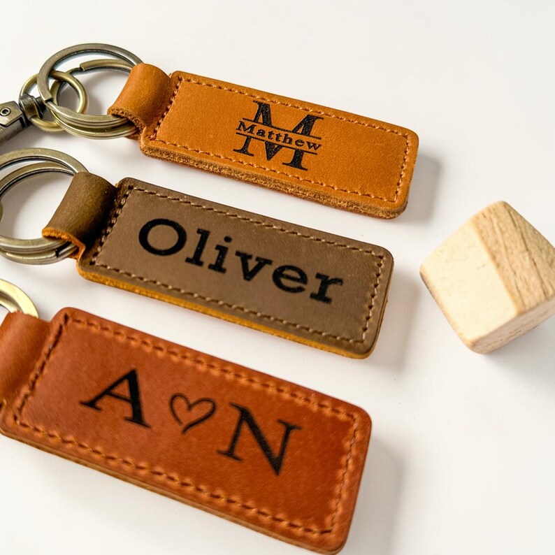May include: Three rectangular leather keychains with metal rings. The top keychain is tan with a black monogram M and the name Matthew. The middle keychain is olive green with the name Oliver. The bottom keychain is brown with the letters A heart N.