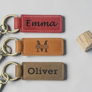 May include: Three rectangular leather keychains with metal rings. The top keychain is red with the name Emma engraved. The middle keychain is tan with the name Matthew and a monogram engraved. The bottom keychain is brown with the name Oliver engraved.