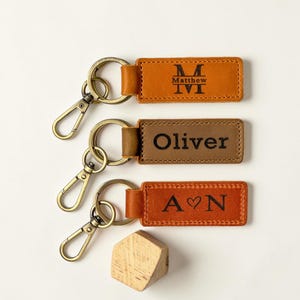 May include: Three rectangular leather keychains with antique brass-toned metal lobster clasps and split rings. The top keychain is tan with the name Matthew and a large M. The middle is brown with the name Oliver. The bottom is orange with A heart N.