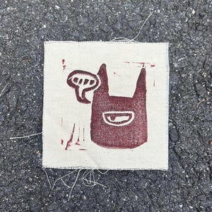 May include: A square, off-white fabric patch featuring a maroon illustration of a cartoon dog with a speech bubble. The dog has pointed ears and a smiling mouth. The patch has frayed edges.