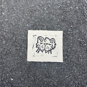 May include: A square, off-white fabric patch featuring a black linocut print of two stylized lion faces. The patch is placed on a dark gray asphalt surface. The lion faces are side-by-side, with simple, bold lines.