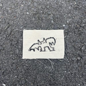 May include: A small, rectangular, off-white fabric patch with a black, stylized illustration of an animal. The animal appears to be a bear or similar creature. The patch has frayed edges and is set against a dark asphalt background.