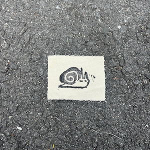 May include: A small, rectangular patch featuring a black snail illustration. The snail is drawn in a simple, cartoon style, with a spiraled shell and a smiling face. The patch is made of a light-coloured fabric.