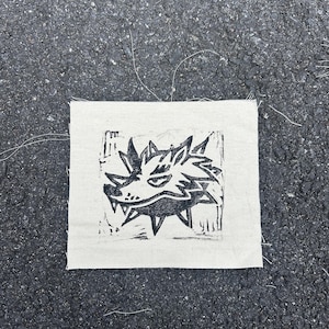 Puede incluir: Un parche de tela cuadrado, blanquecino, con una impresión de linograbado negro de una cabeza de lobo estilizada. El lobo tiene dientes afilados y orejas puntiagudas. La tela tiene bordes sin rematar e hilos visibles.