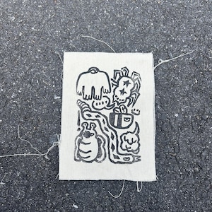 Puede incluir: Un parche de tela cuadrado de color blanquecino con una ilustración negra y caprichosa. El diseño presenta una variedad de figuras de dibujos animados, incluyendo una camisa, una calavera, un insecto y otras formas abstractas. Los bordes del parche están deshilachados.