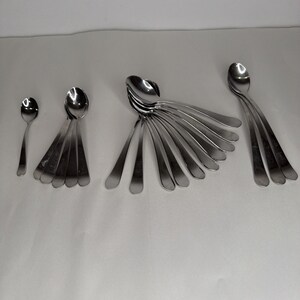 May include: A collection of stainless steel spoons of varying sizes, arranged on a white surface. The spoons have a polished, reflective finish, with elongated handles and rounded bowls. The arrangement includes a single small spoon and several sets of spoons.