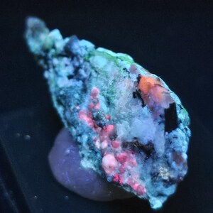 May include: A raw mineral specimen displaying fluorescent properties under ultraviolet light, featuring a textured surface with vibrant patches of neon green, bright pink, and orange crystalline formations against a dark, rocky matrix base.