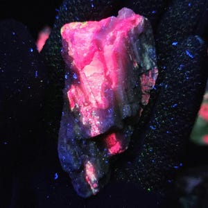 May include: A close-up of a glowing mineral specimen, showcasing vibrant pink and red hues. The rock's surface has a rough texture, with dark areas contrasting the bright colors. Small blue specks are scattered around the mineral, creating a striking visual effect.