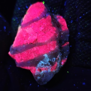 May include: A glowing, irregularly shaped rock with vibrant pink and blue hues. The rock's surface displays a layered pattern with bands of bright pink and areas of blue and white. The background is dark, with scattered blue and green specks.