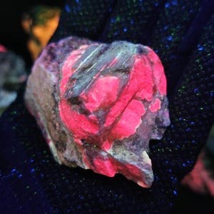 May include: A close-up of a fluorescent mineral specimen. The rock has a rough, textured surface with vibrant pink and gray patches. The background is dark, with scattered blue and green specks.