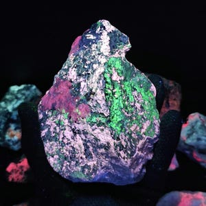 May include: A close-up of a fluorescent mineral specimen. The rock shows a mix of colours, including vibrant green, pink, and blue hues, with a textured surface. The specimen is set against a dark background, highlighting its unique coloration.
