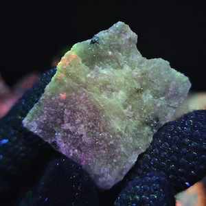 May include: A roughly hewn rock fragment with an irregular shape. The stone displays a pale green and purple colour, with touches of orange and blue. The surface is textured and uneven, with small dark specks. The background is dark.