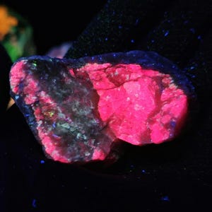 May include: A close-up of a glowing rock with a vibrant pink hue on one side and a darker, speckled appearance on the other. The rock is set against a dark background, with hints of blue and green.